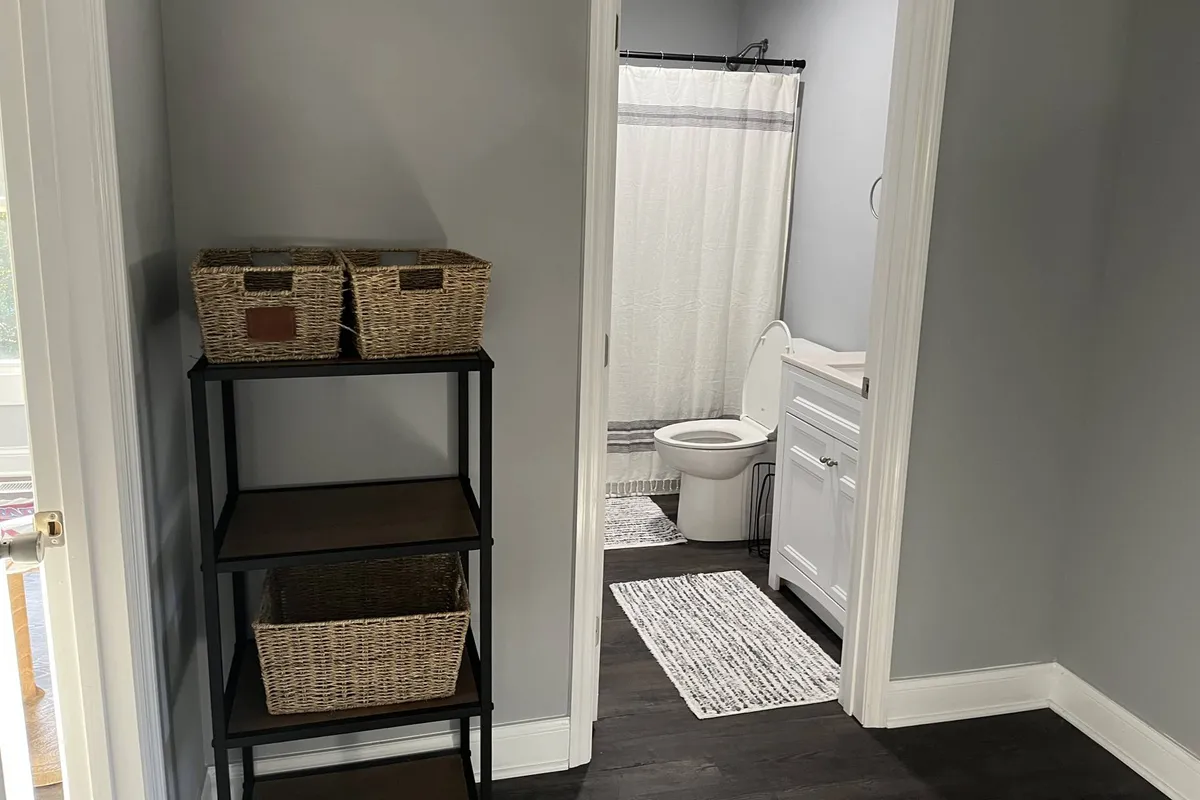 An interior shot showing organized shelves with baskets, a bathroom doorway, and sparkling white bathroom fixtures. The space is filled with calm morning light, suggesting a clean and serene bathroom experience.