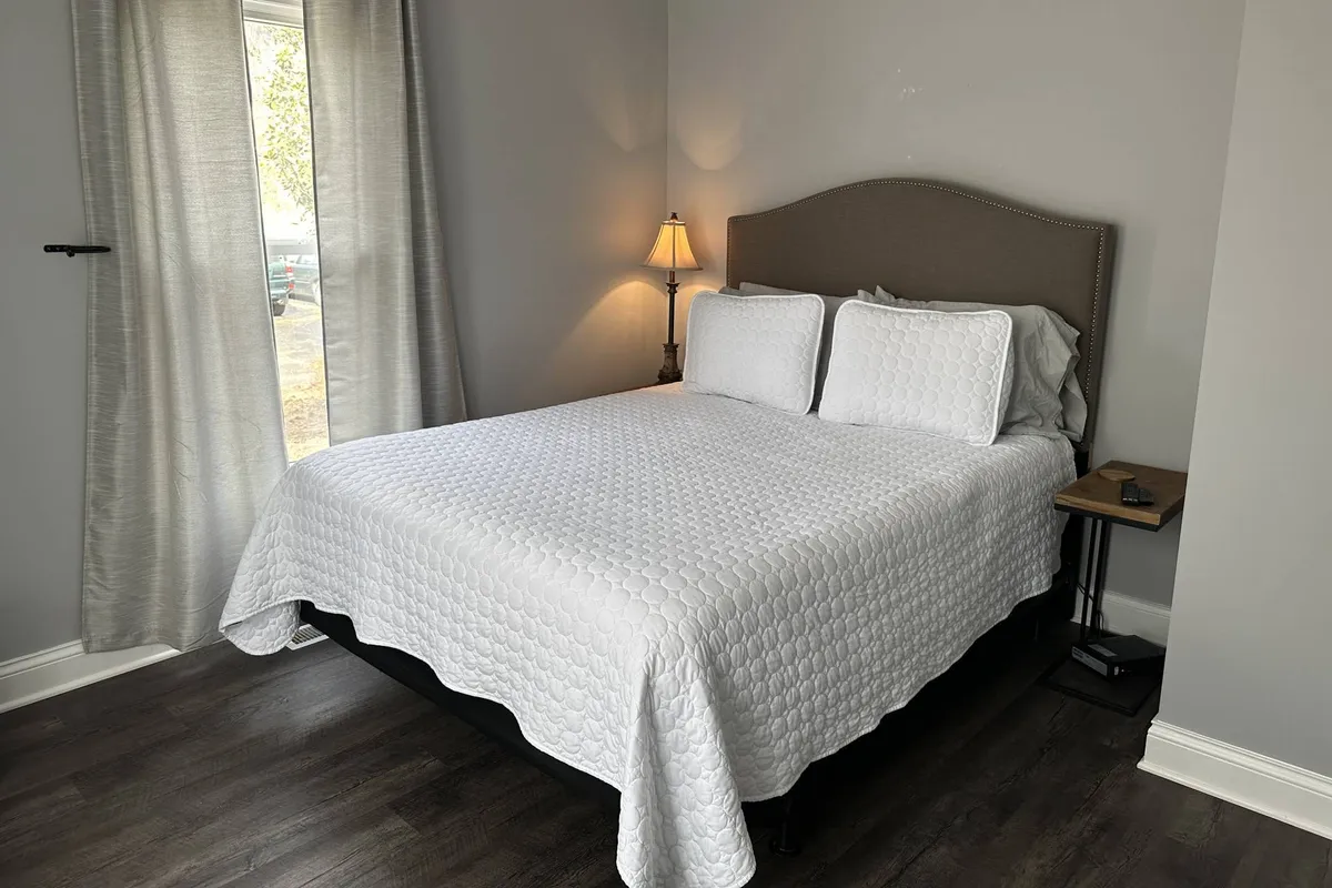 A serene bedroom scene with a neatly made bed, soft lamp lighting, and gentle curtains framing a window. The room is designed for rest and relaxation, with cozy touches that invite a peaceful night's sleep or a quiet moment to read.