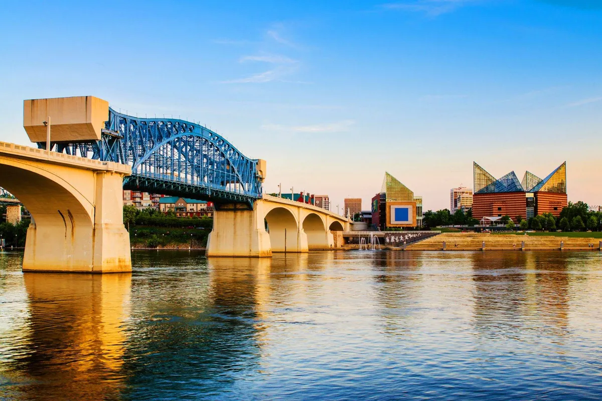 A serene scene of a blue bridge spanning a calm river, flanked by modern buildings and wide riverwalk. The setting sun casts a warm glow over the water, inviting walking, sightseeing, and photography along the riverfront.