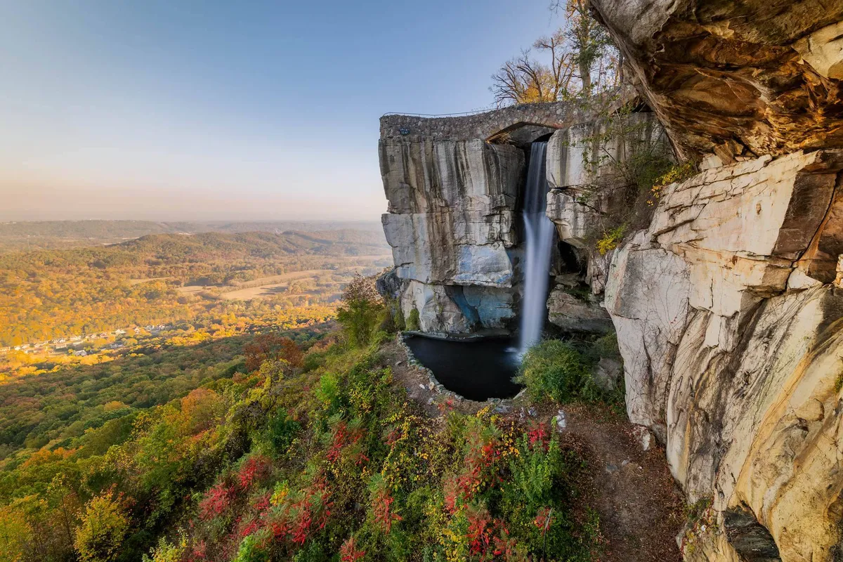 A stunning natural landscape featuring a cascading waterfall amidst dramatic rock formations and a sweeping valley view. Autumn foliage adds vibrant color, and the tranquil setting invites hiking, picnicking, and nature observation.