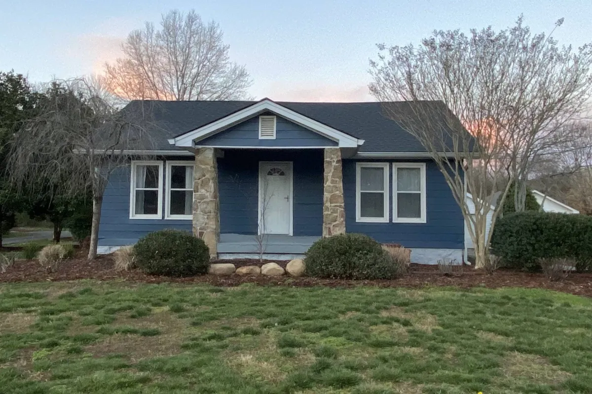 The image showcases the charming blue exterior of a single-family house with inviting stone pillars framing the front door, a well-kept landscaped garden bursting with seasonal blooms, and a welcoming porch ideal for relaxing while watching the sunset. The overall mood is warm and inviting, perfect for family gatherings or outdoor relaxation.
