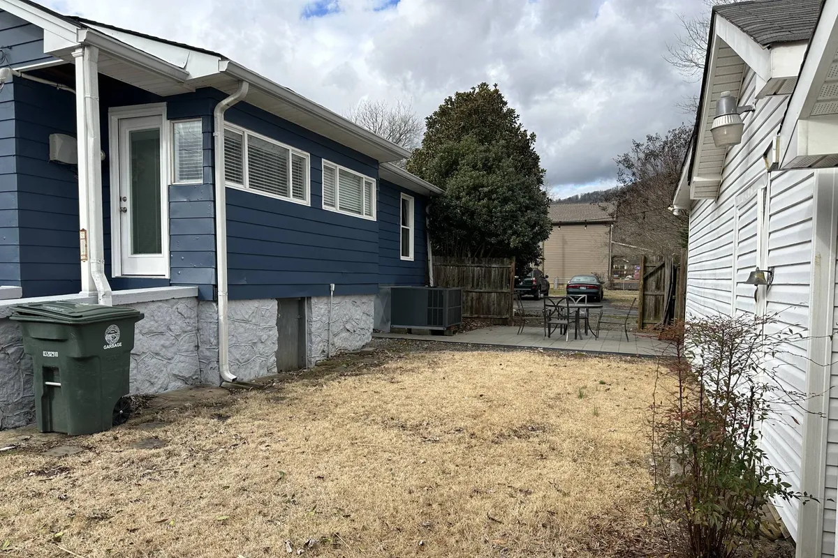 A serene exterior view of a blue house with a cozy patio area, surrounded by a relaxed fenced yard. The patio is set up for outdoor dining or relaxation, emphasizing a sunny and welcoming atmosphere ideal for gatherings or weekend brunches.