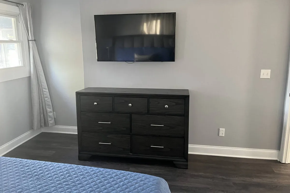 This image features a contemporary bedroom with a bed dressed in blue bedding, a dresser with drawers, and a wall-mounted TV. Natural light filters in through a window, creating a cozy and inviting atmosphere ideal for relaxation or watching TV in bed.