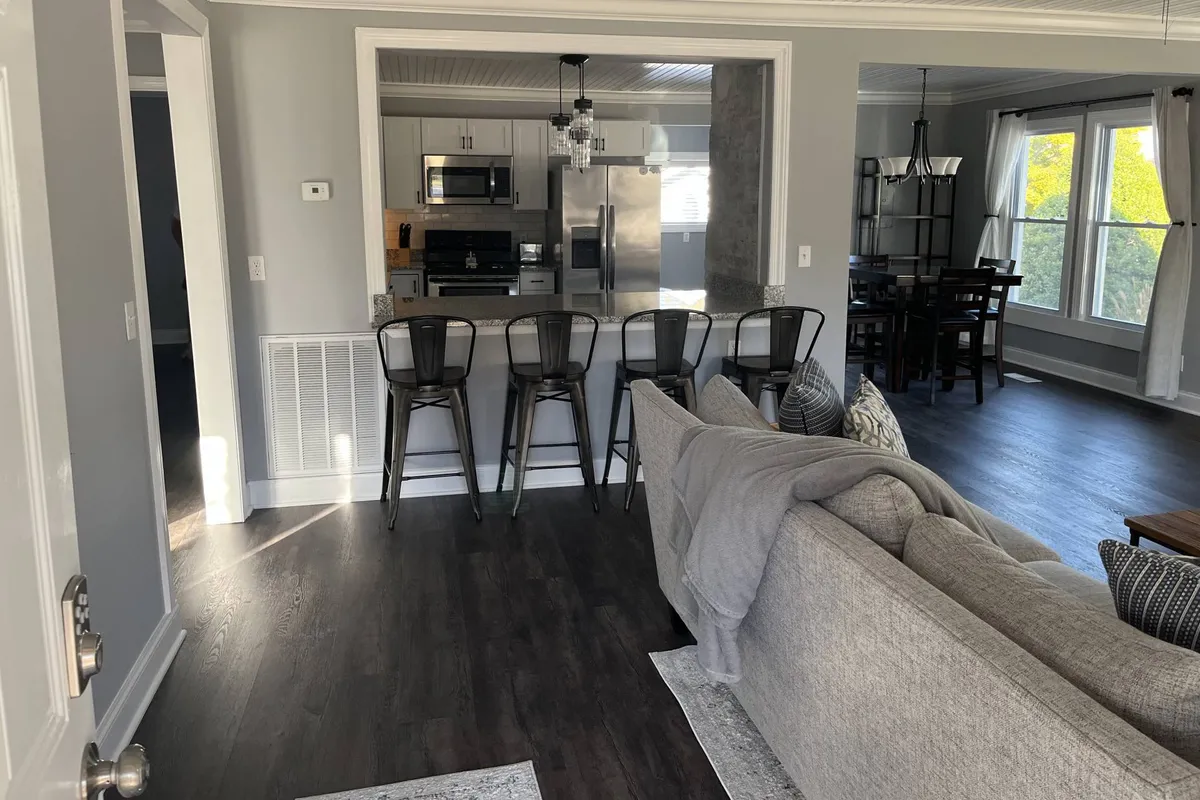 A wide view of the open-concept main living area, showing the kitchen island, a cozy sofa, and the dining table—all seamlessly connected in a single inviting space. Sunlight brightens the room, and seating arrangements suggest a spot for socializing, family gatherings, or quiet relaxation.