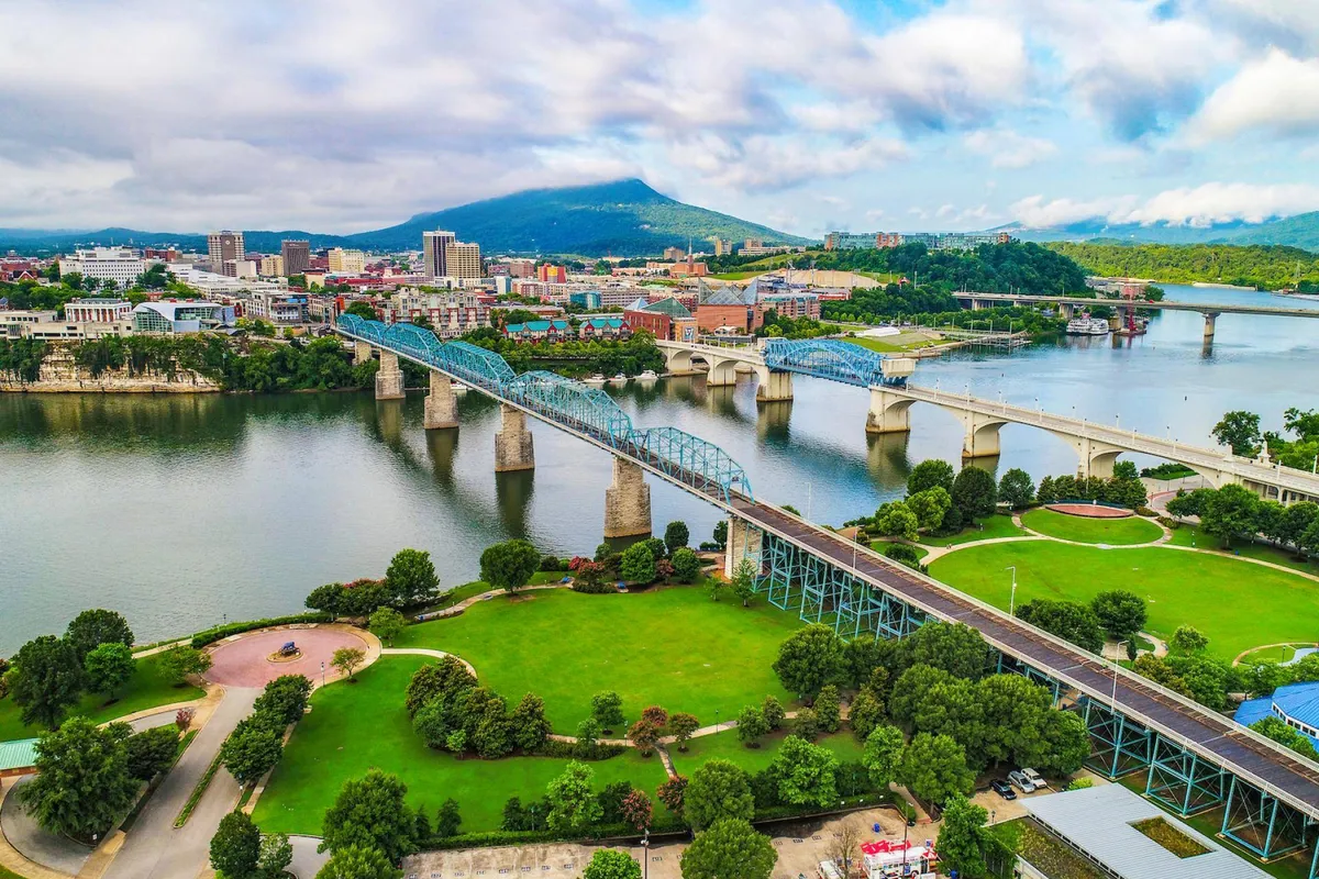 A sweeping view of the Chattanooga riverfront with the sparkling river, city skyline, bridges, green parks, and a backdrop of distant mountains. People are visible walking, cycling, and enjoying the riverside, giving a sense of lively city energy and outdoor activities.