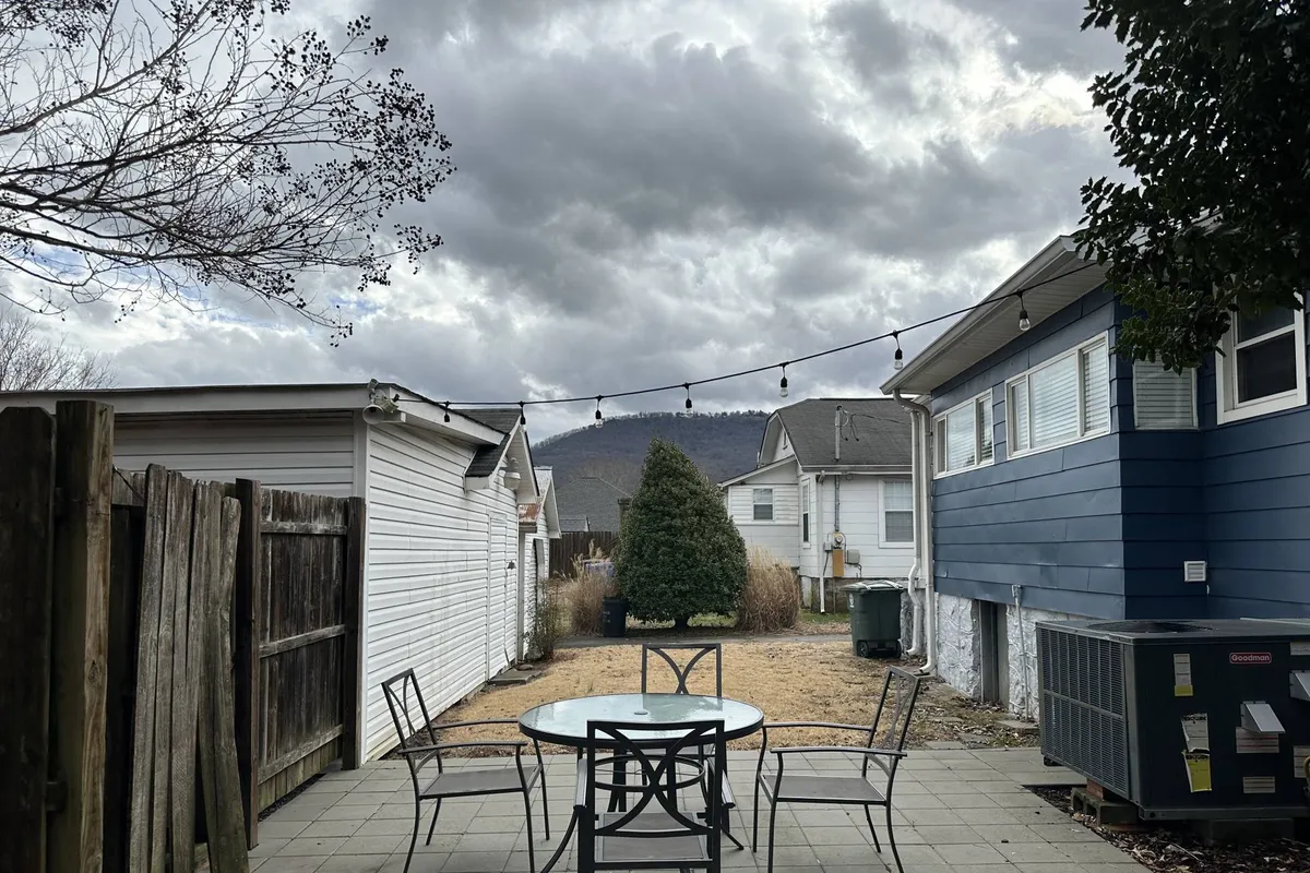 An inviting outdoor dining setup featuring a table and chairs on a deck or patio, illuminated by string lights. In the distance, a breathtaking mountain view sets the scene, and the soft light suggests either early morning or sunset. The overall mood is tranquil and social, perfect for enjoying meals or conversations outdoors.