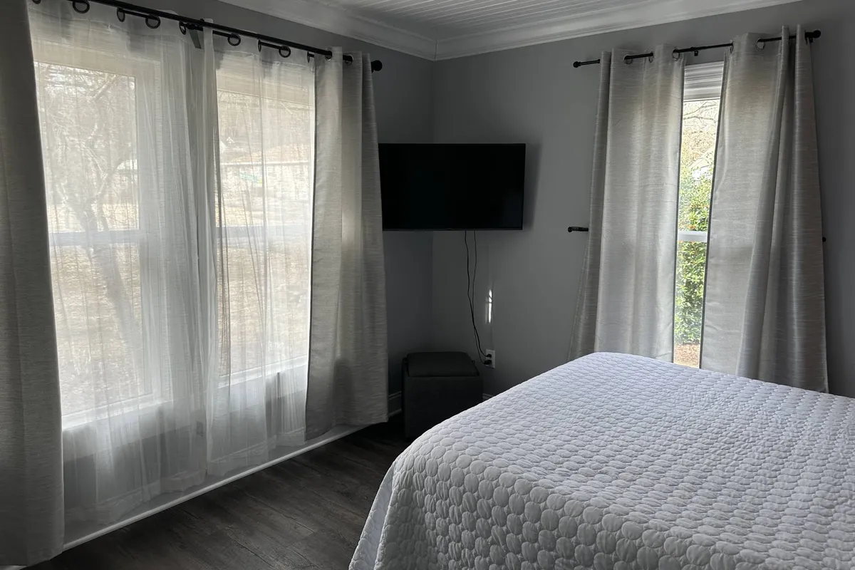 A cozy bedroom with a quilted bed, sheer curtains letting in natural light, and a mounted television at the foot of the bed. The atmosphere is both restful and functional, suitable for unwinding after a busy day or catching up on entertainment.